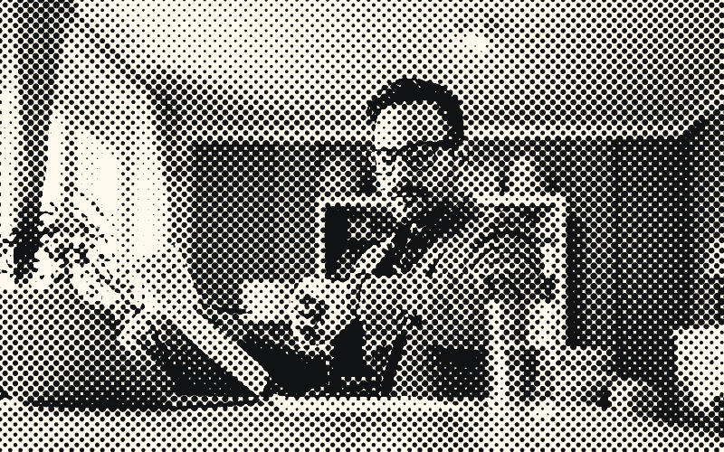 A man in glasses checks his watch at a table, holding a document, with a laptop open in front of him