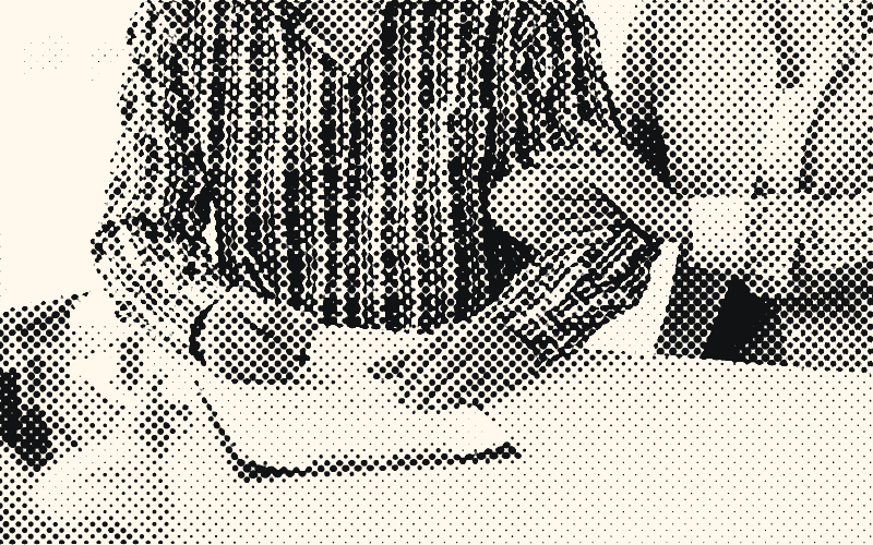 A man in a plaid shirt signs a form on a clipboard while a woman holds his arm; a small house model sits on the desk